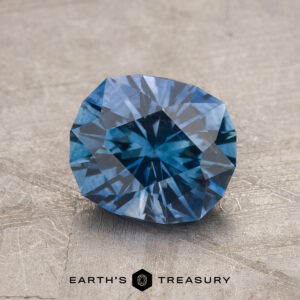 A 1.89-carat Medium Blue Montana Sapphire (Heated) rests on a textured gray surface, with "Earth's Treasury" and a hexagon logo displayed beneath it.