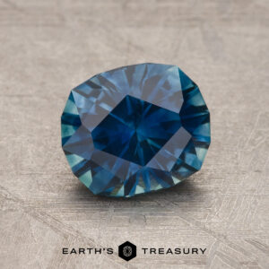 A 1.81-carat Deep Peacock Blue Montana Sapphire (heated) with a cushion cut is displayed on a textured gray surface, with the "Earth's Treasury" logo and name at the bottom of the image.