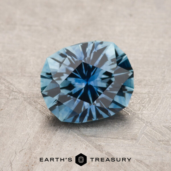 1.65-Carat Medium Blue Montana Sapphire (Heated) A 1.65-Carat Medium Blue Montana Sapphire (Heated) is shown in a round cut on a textured gray surface with "Earth's Treasury" at the bottom.