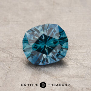 A 1.35-Carat Rich Teal Montana Sapphire (Heated) with geometric facets rests on a textured gray surface, "Earth's Treasury" displayed below the gem.