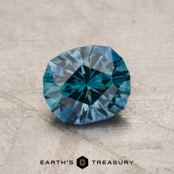 1.35-Carat Rich Teal Montana Sapphire (Heated) A 1.35-Carat Rich Teal Montana Sapphire (Heated) with geometric facets rests on a textured gray surface, "Earth's Treasury" displayed below the gem.