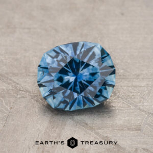 A 1.27-carat medium blue Montana Sapphire (heated) is displayed on a textured surface with "Earth's Treasury" text and logo below.