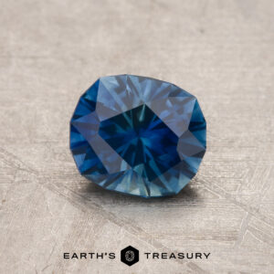 A 1.13-carat blue-teal particolored Montana Sapphire (heated) rests on a textured surface, with the "Earth's Treasury" logo and text at the bottom.