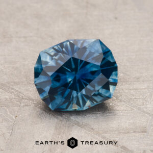 A faceted 1.52-Carat Peacock Blue Montana Sapphire (Heated) is shown on a light textured surface, with the "Earth's Treasury" text and logo beneath it.