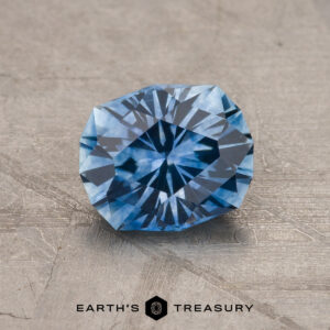 A 1.43-carat medium blue heated Montana Sapphire with a geometric faceted pattern rests on a textured gray surface. "EARTH'S TREASURY" is displayed at the bottom.