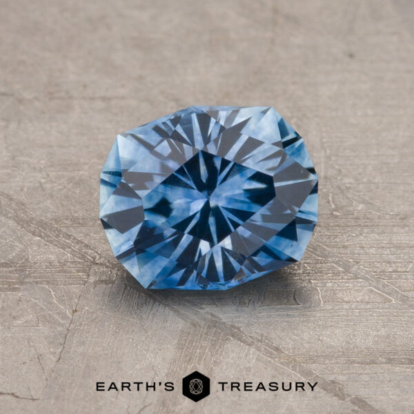 1.43-Carat Medium Blue Montana Sapphire (Heated) A 1.43-carat medium blue heated Montana Sapphire with a geometric faceted pattern rests on a textured gray surface. "EARTH'S TREASURY" is displayed at the bottom.