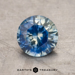 A 1.76-carat blue-yellow particolored heated Montana Sapphire gemstone displayed on a textured gray surface, with the Earth's Treasury logo beneath.