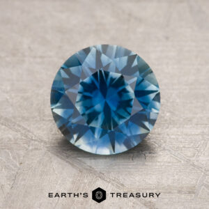 A 1.61-carat rich teal blue Montana sapphire (heated), round brilliant-cut, is displayed on a textured surface with the "Earth's Treasury" logo and text at the bottom.