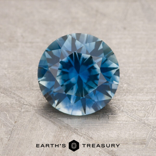 1.61-Carat Rich Teal Blue Montana Sapphire (Heated) A 1.61-carat rich teal blue Montana sapphire (heated), round brilliant-cut, is displayed on a textured surface with the "Earth's Treasury" logo and text at the bottom.