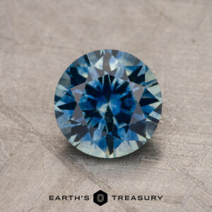 A 1.44-carat teal blue Montana sapphire (heated), round-cut with a faceted surface, is displayed on a textured gray background. The text "EARTH'S TREASURY" appears below the gem.