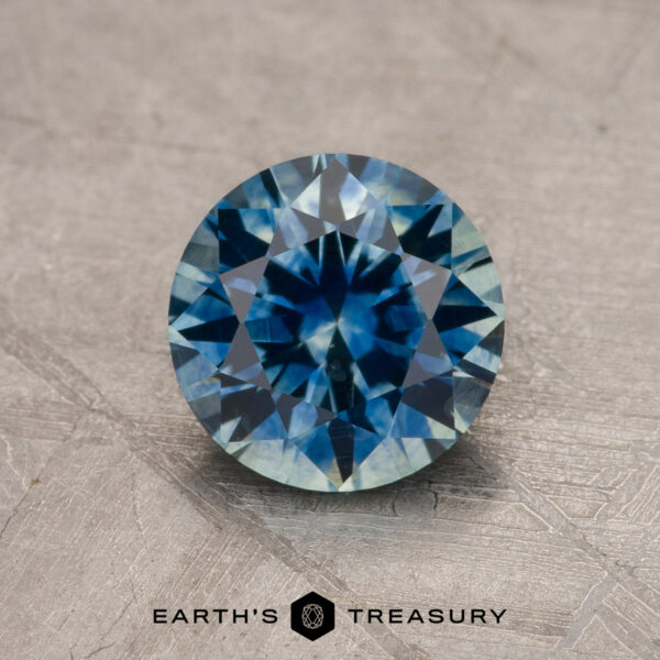 1.44-Carat Teal Blue Montana Sapphire (Heated) A 1.44-carat teal blue Montana sapphire (heated), round-cut with a faceted surface, is displayed on a textured gray background. The text "EARTH'S TREASURY" appears below the gem.