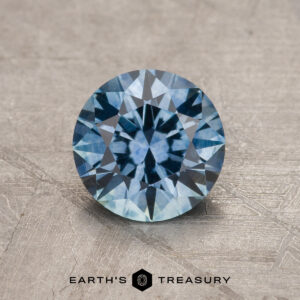 A 1.25-Carat Sky Blue Montana Sapphire (Heated) is shown in a round brilliant cut on a textured gray surface with the Earth's Treasury logo and text below.