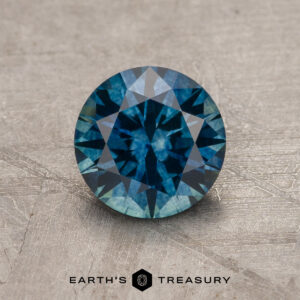 1.24-Carat Rich Teal Montana Sapphire (Heated), round brilliant-cut, displayed on a textured light-colored surface with the "Earth's Treasury" logo and text at the bottom.