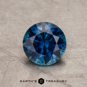 A 1.21-Carat Deep Peacock Blue Montana Sapphire (Heated), round brilliant-cut, rests on a textured surface with the "Earth's Treasury" logo and text displayed at the bottom of the image.