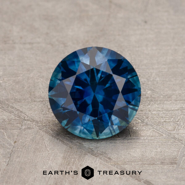1.21-Carat Deep Peacock Blue Montana Sapphire (Heated) A 1.21-Carat Deep Peacock Blue Montana Sapphire (Heated), round brilliant-cut, rests on a textured surface with the "Earth's Treasury" logo and text displayed at the bottom of the image.