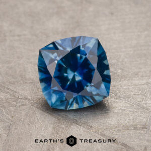 A 1.23-carat rich blue heated Montana Sapphire with a cushion cut and intricate facets is displayed on a light textured surface. "EARTH'S TREASURY" appears at the bottom of the image.