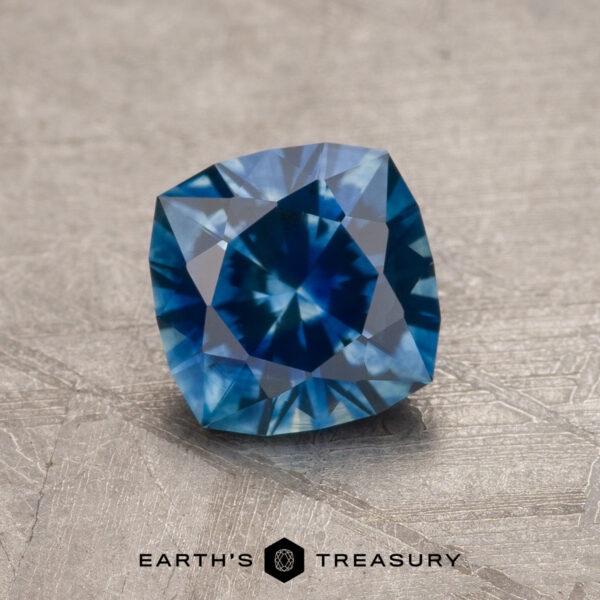 1.23-Carat Rich Blue Montana Sapphire (Heated) A 1.23-carat rich blue heated Montana Sapphire with a cushion cut and intricate facets is displayed on a light textured surface. "EARTH'S TREASURY" appears at the bottom of the image.
