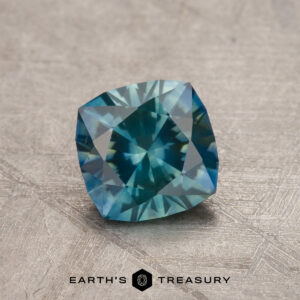 A 1.12-Carat Teal Montana Sapphire (Heated) is displayed on a textured surface with the "Earth's Treasury" logo beneath it.