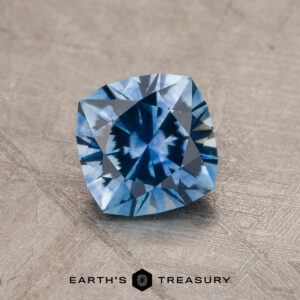 A 1.11-carat medium blue Montana Sapphire (heated) with a square cushion cut rests on a textured gray surface. The "Earth's Treasury" logo is visible at the bottom of the image.
