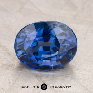 A 5.21-Carat Sapphire, expertly cut in an oval shape, is showcased on a light textured surface with the "Earth's Treasury" logo and text displayed below.