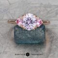 The "Lilium" ring in 14k rose gold, a stunning piece of jewelry, showcases a 1.72-carat Montana sapphire as its central feature, complemented by two delicate pink stones. Perfect for fashion enthusiasts, it is displayed elegantly on a textured surface.
