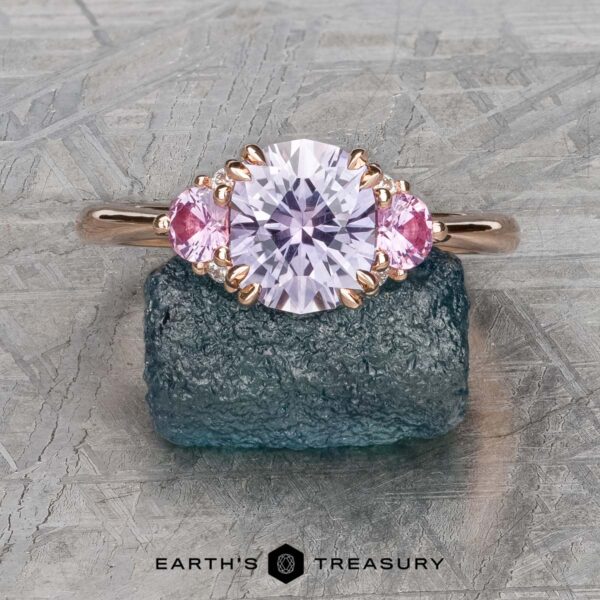The "Lilium" ring in 14k rose gold with 1.72-Carat Montana Sapphire The "Lilium" ring in 14k rose gold, a stunning piece of jewelry, showcases a 1.72-carat Montana sapphire as its central feature, complemented by two delicate pink stones. Perfect for fashion enthusiasts, it is displayed elegantly on a textured surface.