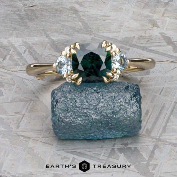 The "Lillium" ring in 14k yellow gold with 1.38-carat Australian sapphire The "Lillium" ring in 14k yellow gold with a 1.38-carat Australian sapphire, accented by two clear stones and shown on a rough blue crystal, with the Earth's Treasury logo beneath.