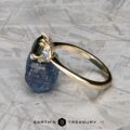 The "Lillium" ring in 14k yellow gold with a 1.38-carat Australian sapphire, accented by two clear stones and shown on a rough blue crystal, with the Earth's Treasury logo beneath.