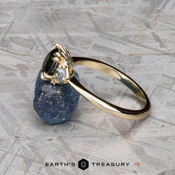 The "Lillium" ring in 14k yellow gold with 1.38-carat Australian sapphire The "Lillium" ring in 14k yellow gold with a 1.38-carat Australian sapphire, accented by two clear stones and shown on a rough blue crystal, with the Earth's Treasury logo beneath.
