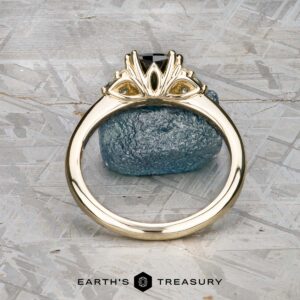 The "Lillium" ring in 14k yellow gold with a 1.38-carat Australian sapphire, accented by two clear stones and shown on a rough blue crystal, with the Earth's Treasury logo beneath.