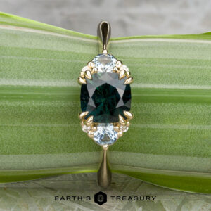 The "Lillium" ring in 14k yellow gold, featuring a 1.38-carat Australian sapphire and two light blue stones, is shown on a green leaf background with the "Earth's Treasury" logo below.