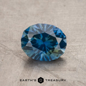 A 1.32-carat blue-yellow particolored Montana sapphire (heated), oval-cut with intricate facets, rests on a textured surface; the "Earth's Treasury" logo appears below the gem.
