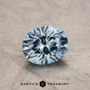 A 1.28-carat aqua blue Montana sapphire (heated), round brilliant-cut, shines on a textured gray surface with "Earth's Treasury" displayed below.