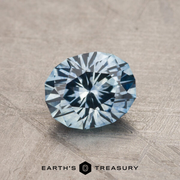 1.28-Carat Aqua Blue Montana Sapphire (Heated) A 1.28-carat aqua blue Montana sapphire (heated), round brilliant-cut, shines on a textured gray surface with "Earth's Treasury" displayed below.