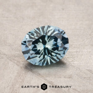 A 1.17-carat aqua blue Montana sapphire (heated), oval faceted, rests on a textured surface with "EARTH'S TREASURY" displayed at the bottom.