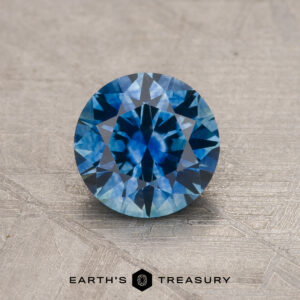 A 1.15-carat medium blue Montana Sapphire (heated), brilliant-cut and round, rests on a textured beige surface with the Earth’s Treasury logo and text displayed below.