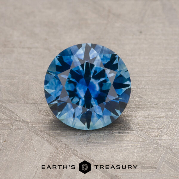 1.15-Carat Medium Blue Montana Sapphire (Heated) A 1.15-carat medium blue Montana Sapphire (heated), brilliant-cut and round, rests on a textured beige surface with the Earth’s Treasury logo and text displayed below.