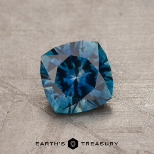 A 1.13-Carat Peacock Blue Montana Sapphire (Heated), cushion-cut, sits on a textured gray surface with the "Earth's Treasury" logo and text beneath it.