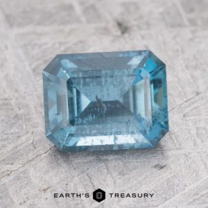 A rectangular, teal gemstone with a faceted cut against a textured, light gray background. Text at the bottom reads "EARTH'S TREASURY: 1.68-Carat Nasarawa Teal Aqua.