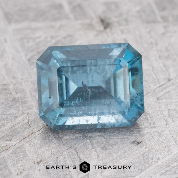 1.68-Carat Nasarawa Teal Aqua A rectangular, teal gemstone with a faceted cut against a textured, light gray background. Text at the bottom reads "EARTH'S TREASURY: 1.68-Carat Nasarawa Teal Aqua.