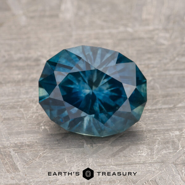 1.93-Carat Deep Blue-Green Montana Sapphire (Heated) A 1.93-carat Montana sapphire (heated), featuring a faceted blue gemstone with a star-like pattern at its center, displayed on a textured surface. The label "Earth's Treasury" is present at the bottom of the image.