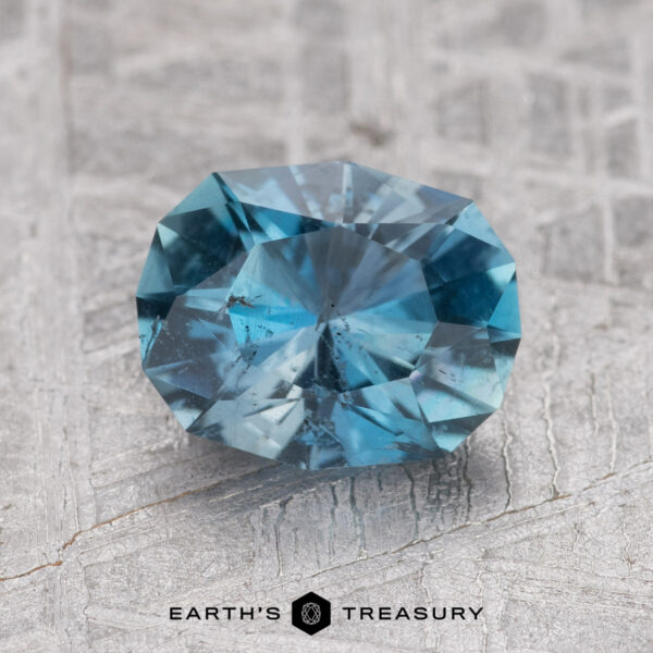 1.38-Carat Nasarawa Teal Aqua A faceted 1.38-carat Nasarawa Teal Aqua gemstone with slight inclusions is placed on a textured, light gray surface. The text "Earth's Treasury" is visible at the bottom of the image.
