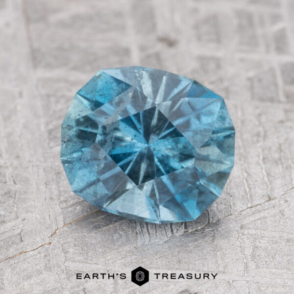 A faceted 1.48-carat Nasarawa Teal Aqua gemstone rests on a textured, gray surface. The text "EARTH'S TREASURY" is printed below the gemstone.
