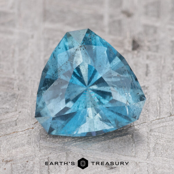 1.67-Carat Nasarawa Teal Aqua A 1.67-carat Nasarawa Teal Aqua gemstone with a triangular shape sits on a textured surface. The caption "Earth's Treasury" is visible at the bottom of the image.