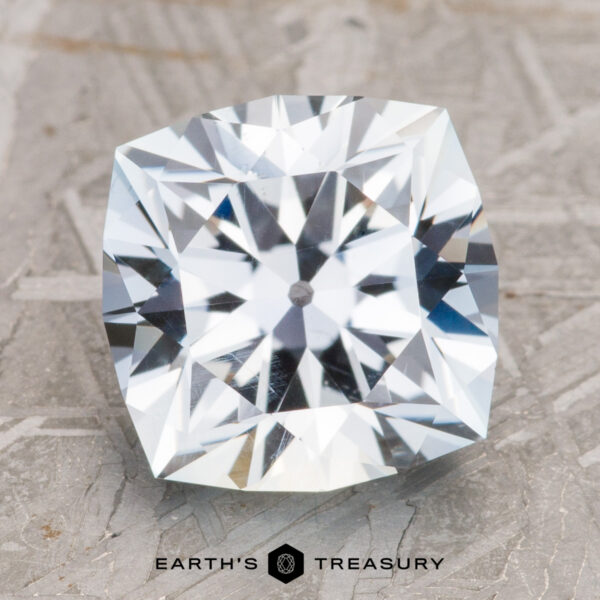 4.23-Carat Pale Aqua Montana Sapphire A close-up photo of the polished, cushion-cut 4.23-Carat Pale Aqua Montana Sapphire, featuring a "Earth's Treasury" logo at the bottom center. The gem exhibits a stunning array of sparkling facets against a textured surface background.