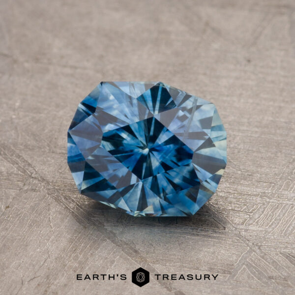 1.79-Carat Montana Sapphire (Heated) A close-up image of a faceted teal blue Montana sapphire, weighing 1.79 carats and featuring intricate cuts, placed on a textured surface. The bottom center of the image features the text "EARTH'S TREASURY" along with a geometric logo.