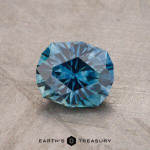 A faceted, teal-blue 1.79-carat Montana sapphire gemstone (heated) rests on a textured gray background. The gemstone is intricately cut with numerous facets, forming a geometric pattern and reflecting light. The text "EARTH'S TREASURY" appears at the bottom of the image.