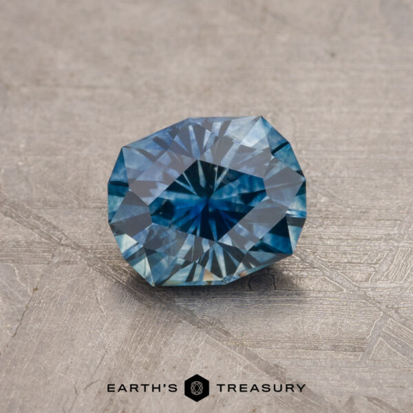 1.17-Carat Montana Sapphire (Heated) A faceted 1.79-carat teal blue Montana sapphire with intricate cuts displayed on a textured, grayish surface. The gem exhibits a mix of dark and light teal blue hues, and the background features a subtle geometric design. "Earth's Treasury" is written at the bottom of the image.