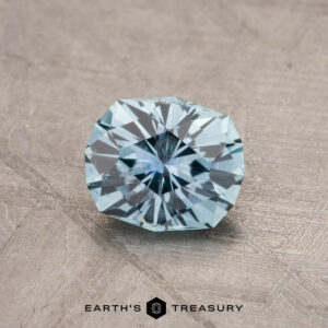 A shiny, intricately faceted 1.79-carat teal blue Montana sapphire is placed on a textured, gray surface. The gemstone has sharp edges and reflects light, showcasing its clarity and cut. At the bottom, the text "EARTH'S TREASURY" is visible in black letters.