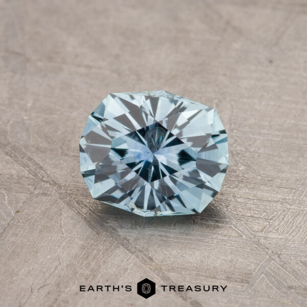 1.12-Carat Montana Sapphire (Heated) A shiny, intricately faceted 1.79-carat teal blue Montana sapphire is placed on a textured, gray surface. The gemstone has sharp edges and reflects light, showcasing its clarity and cut. At the bottom, the text "EARTH'S TREASURY" is visible in black letters.